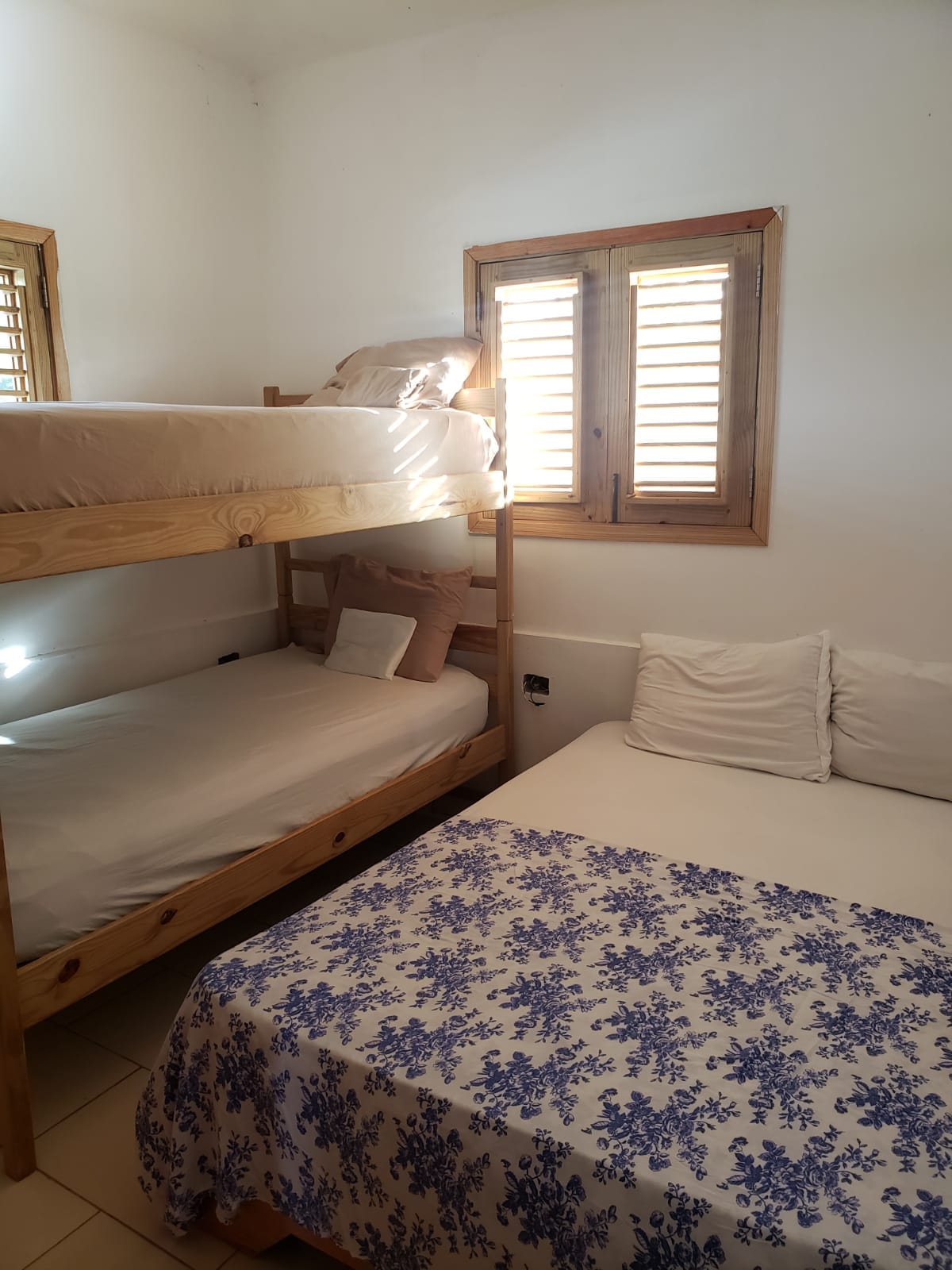 A sleeping area features a wooden bunk bed with a spacious bottom mattress and a soft upper mattress. Light streams through shuttered windows, illuminating the room's neutral walls. A floral-patterned blanket covers the larger bed, which is accompanied by several cushions.