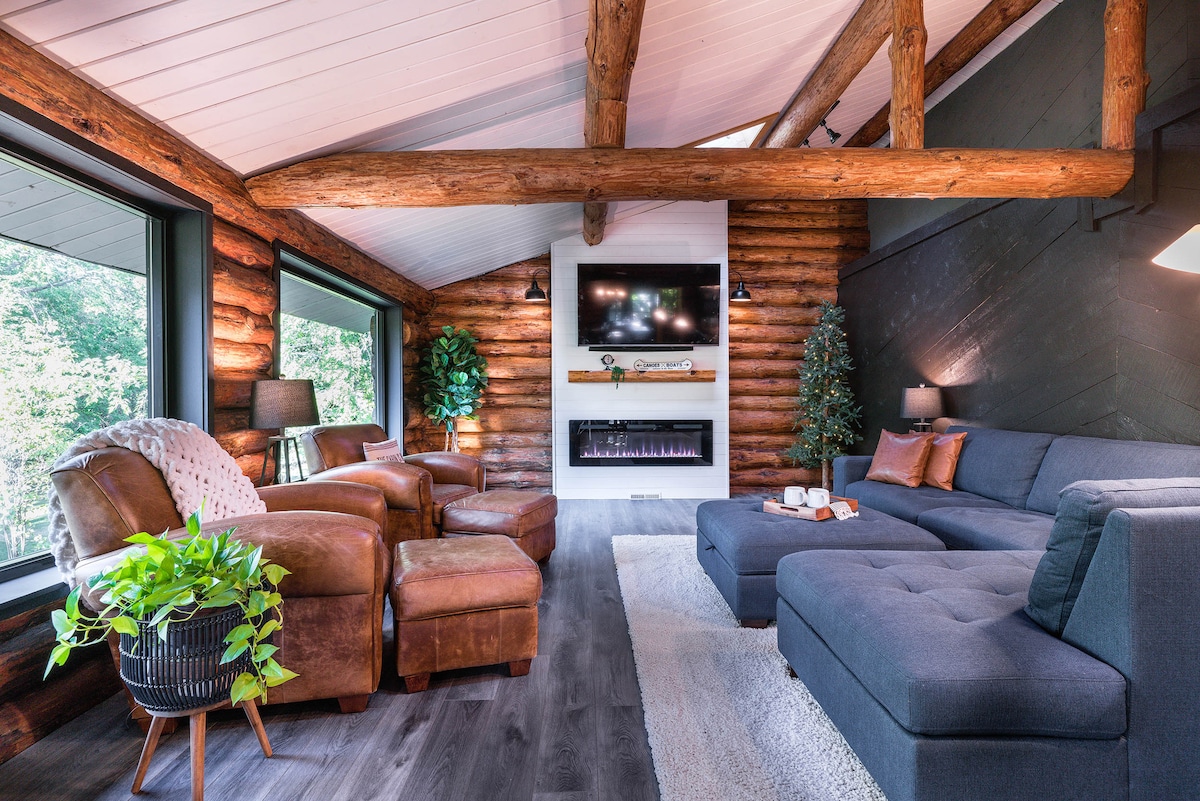 The spacious living area features rustic log walls accented by modern furnishings. A sectional couch is complemented by a leather chair, while a flat-screen TV and electric fireplace occupy the wall. Large windows provide views of the surrounding nature, enhancing the tranquil environment.
