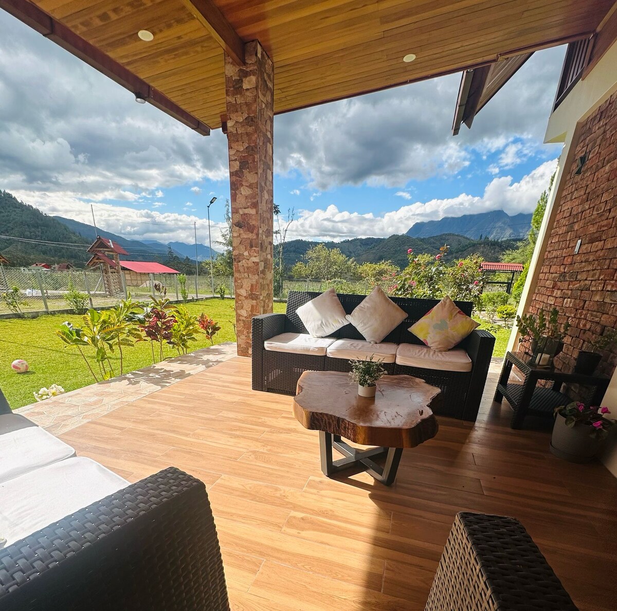 An outdoor seating area features a dark wicker couch adorned with colorful cushions. A natural wood coffee table occupies the center, offering a view of rolling hills and a cloudy sky. Lush green plants and flowers complement the tranquil setting.