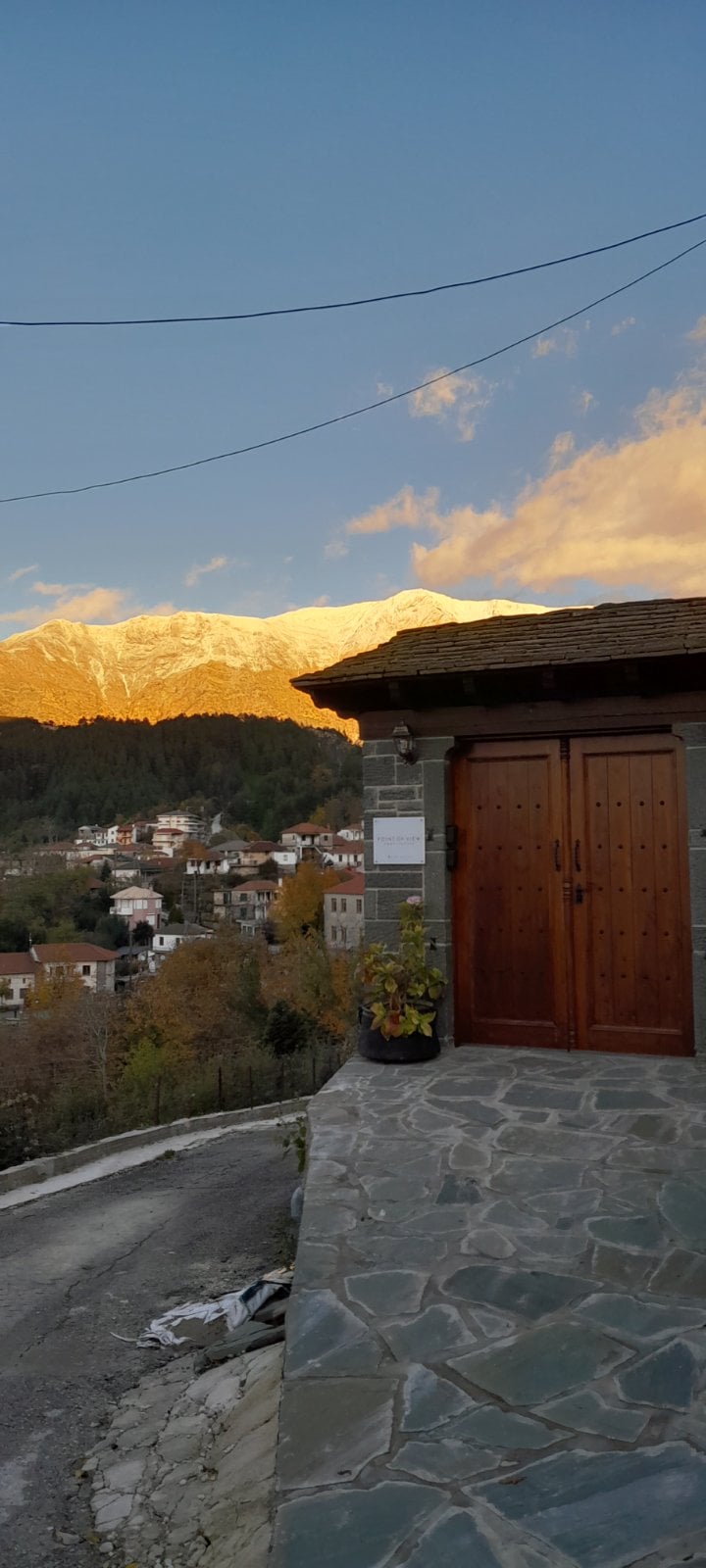 The entrance features sturdy wooden doors framed by a stone facade. A view of the village is visible in the background, with mountains illuminated by the golden light of sunset. A paved path leads up to the entrance, bordered by decorative foliage.