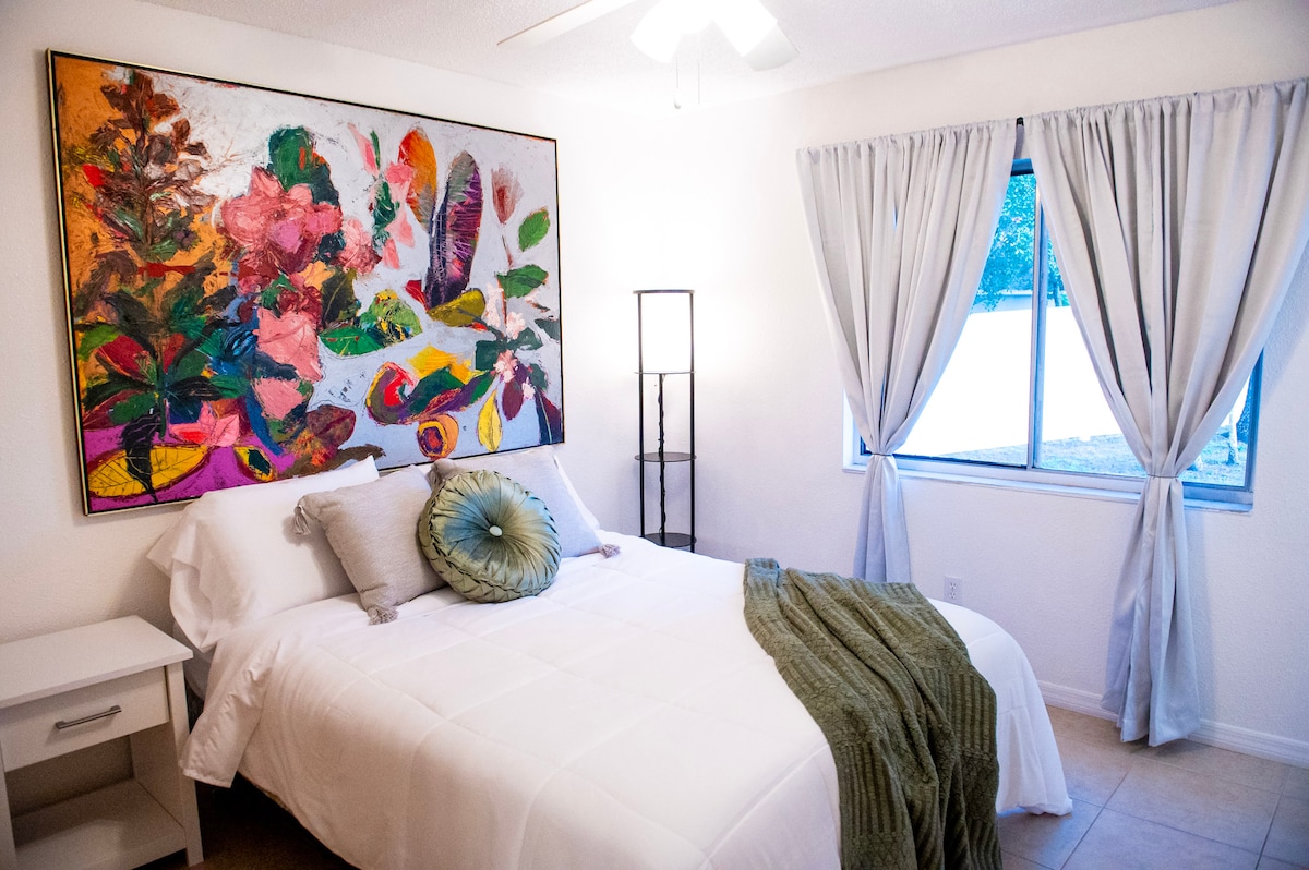 El dormitorio principal ofrece un refugio tranquilo con una cama lujosa, una enorme ventana que inunda el espacio con luz natural y un llamativo cuadro sobre ella, que combina comodidad y creatividad en un entorno de estilo precioso.