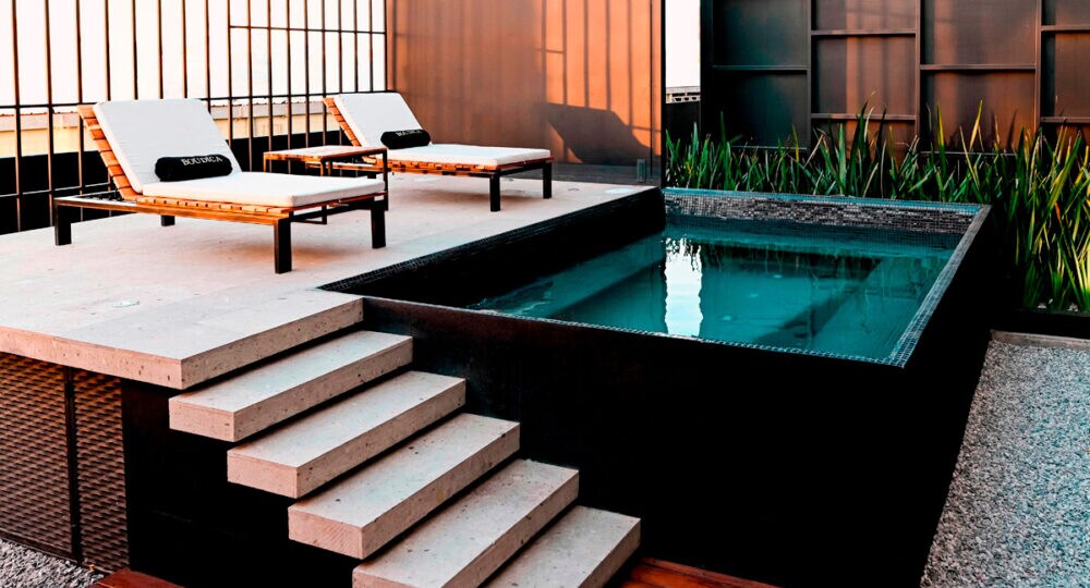 An outdoor space features a sleek jacuzzi, surrounded by stone steps and two wooden lounge chairs. Minimalist design and greenery enhance the modern ambiance, providing a serene environment for relaxation.