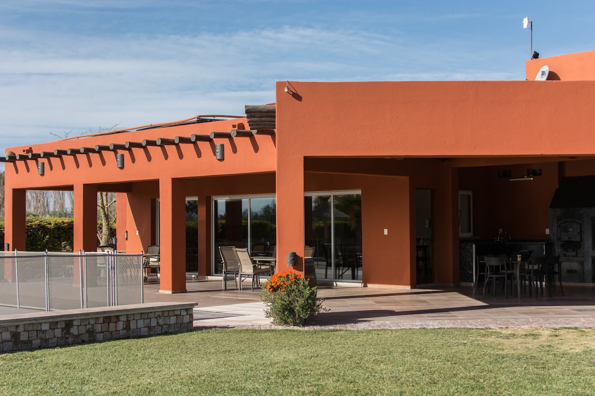 A modern orange structure with large glass sliding doors showcases an inviting outdoor dining area. The spacious patio features a barbecue zone and well-maintained landscaping, with a green lawn and decorative flowers enhancing the outdoor ambiance.