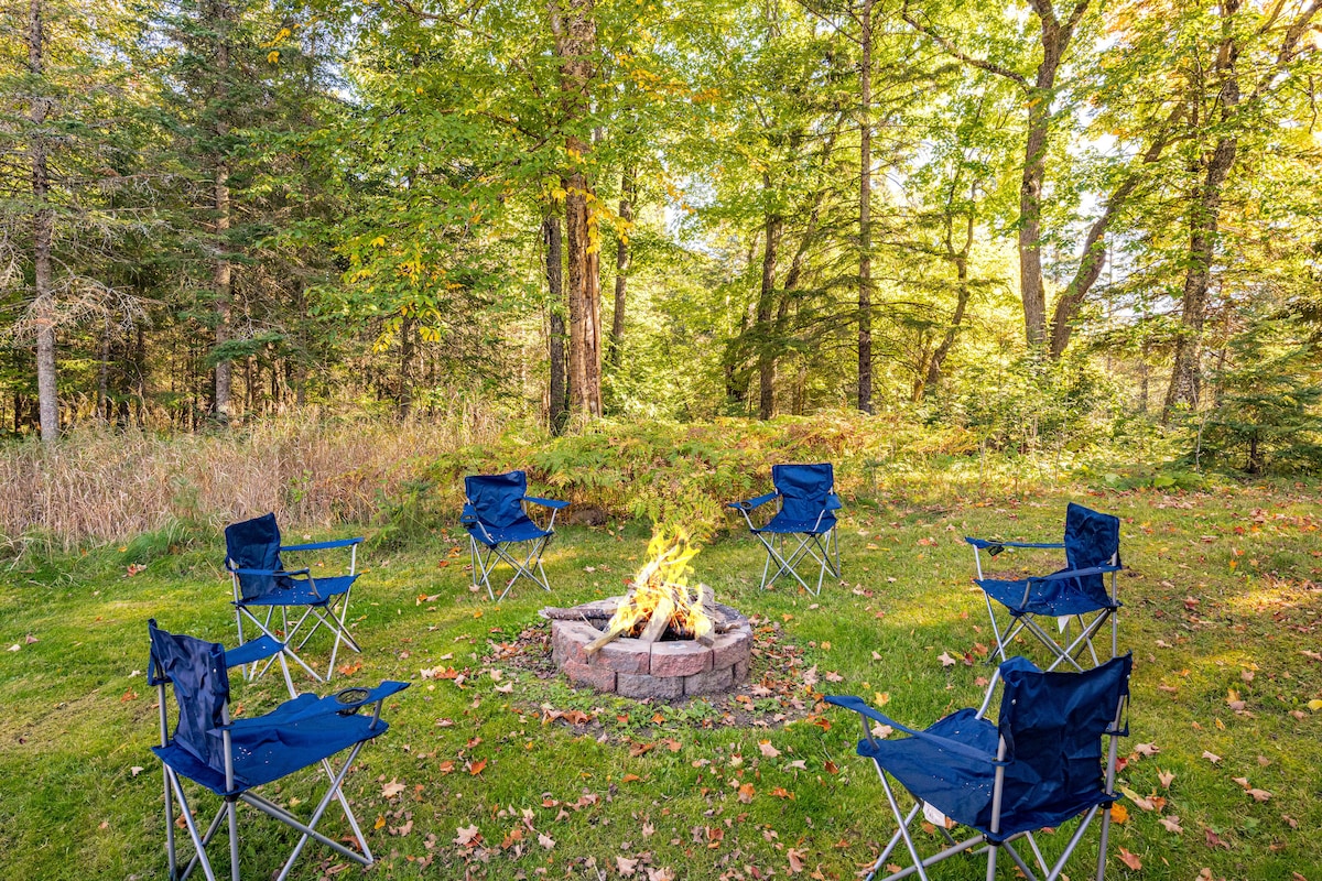 A circular arrangement of blue camping chairs surrounds a stone fire pit, situated in a forested area that features lush green grass and trees. The warm glow of the fire contrasts with the vibrant colors of the surrounding foliage.