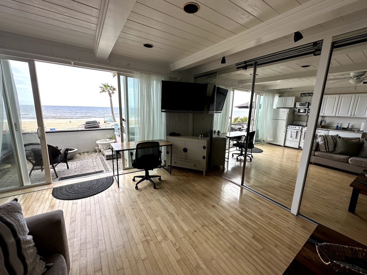 A spacious living area opens to the beach, featuring large glass doors that provide a clear view of the ocean. A table with a chair is positioned alongside a wall-mounted television. The design includes wooden flooring and light curtains that softly diffuse natural light.