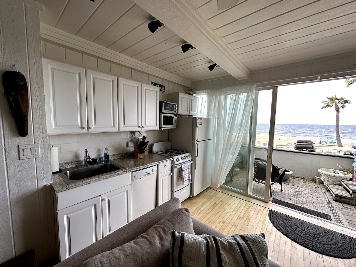 The kitchen area features white cabinetry and granite countertops with a sink and appliances visible, including a microwave and refrigerator. A cozy seating area is situated to the left, and large glass doors open to reveal a view of the beach and palm trees.