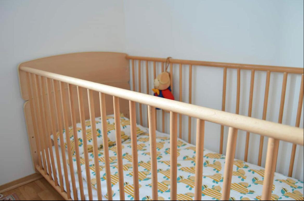 A wooden crib is positioned against a light-colored wall, featuring a soft bedding set decorated with a playful pattern. A small plush toy hangs gently from the crib's side, adding a comforting touch to the space.