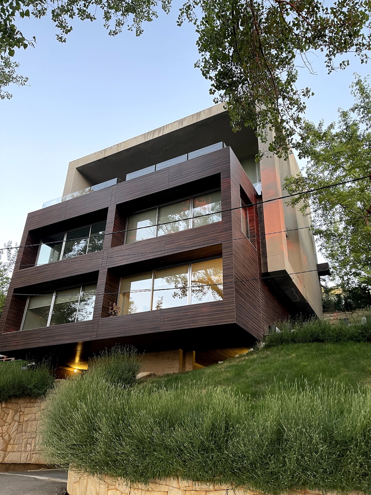 A modern chalet is displayed, characterized by large glass windows that invite natural light. The structure is surrounded by lush greenery, with a slight elevation providing a view of the landscape. The exterior features a mix of wood and concrete for a contemporary look.