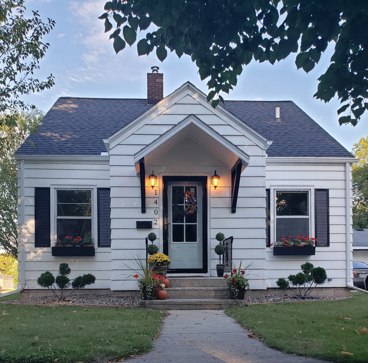 A charming Cape Cod-style cottage is captured from the front, featuring white siding and dark shutters. A welcoming entrance with a decorative wreath is framed by flower boxes filled with colorful blooms. A manicured lawn and a concrete walkway lead up to the door.
