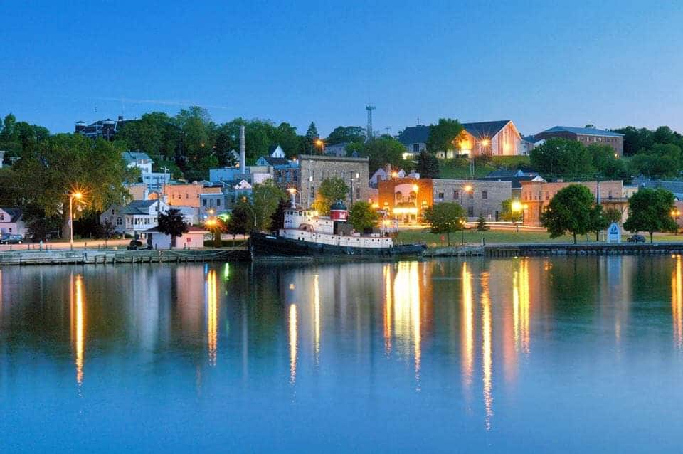 A tranquil evening view of the historic harbor town of Kewaunee is presented. Soft lights reflect off the calm water, highlighting charming buildings along the shore, with trees lining the waterfront. A vintage boat is anchored, adding character to the serene landscape.