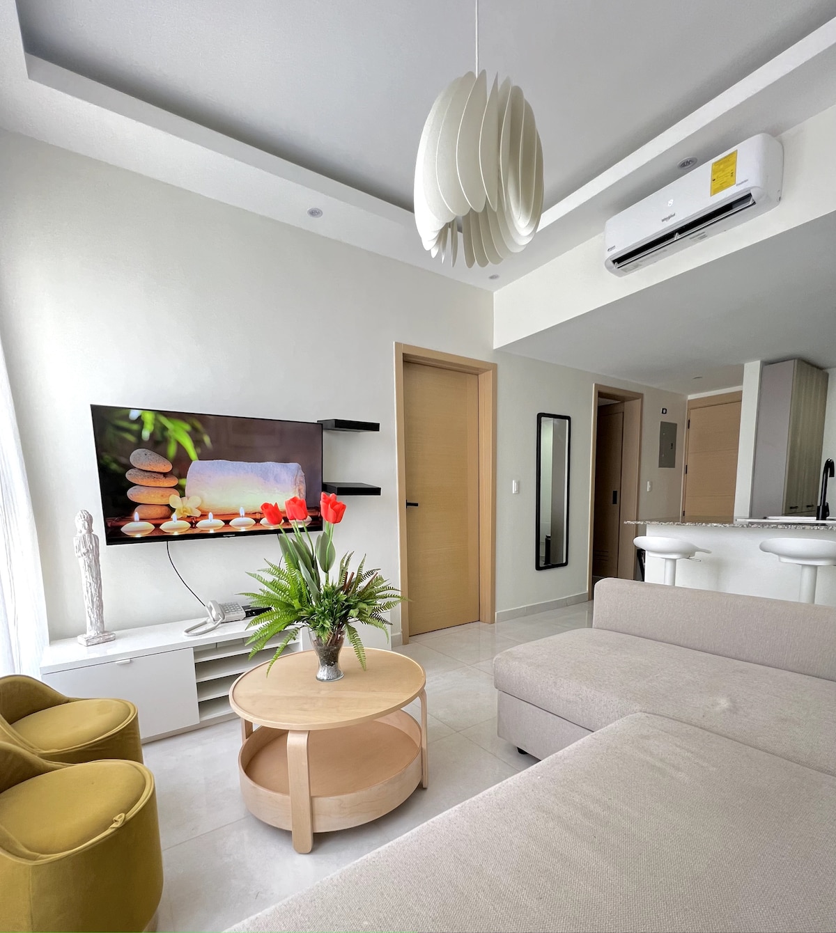 A modern living area features a light-colored sofa paired with a round wooden coffee table. Decorative plants and vibrant flowers are arranged on the table. A flat-screen TV is mounted on the wall, providing access to entertainment. Large windows allow natural light to fill the space.