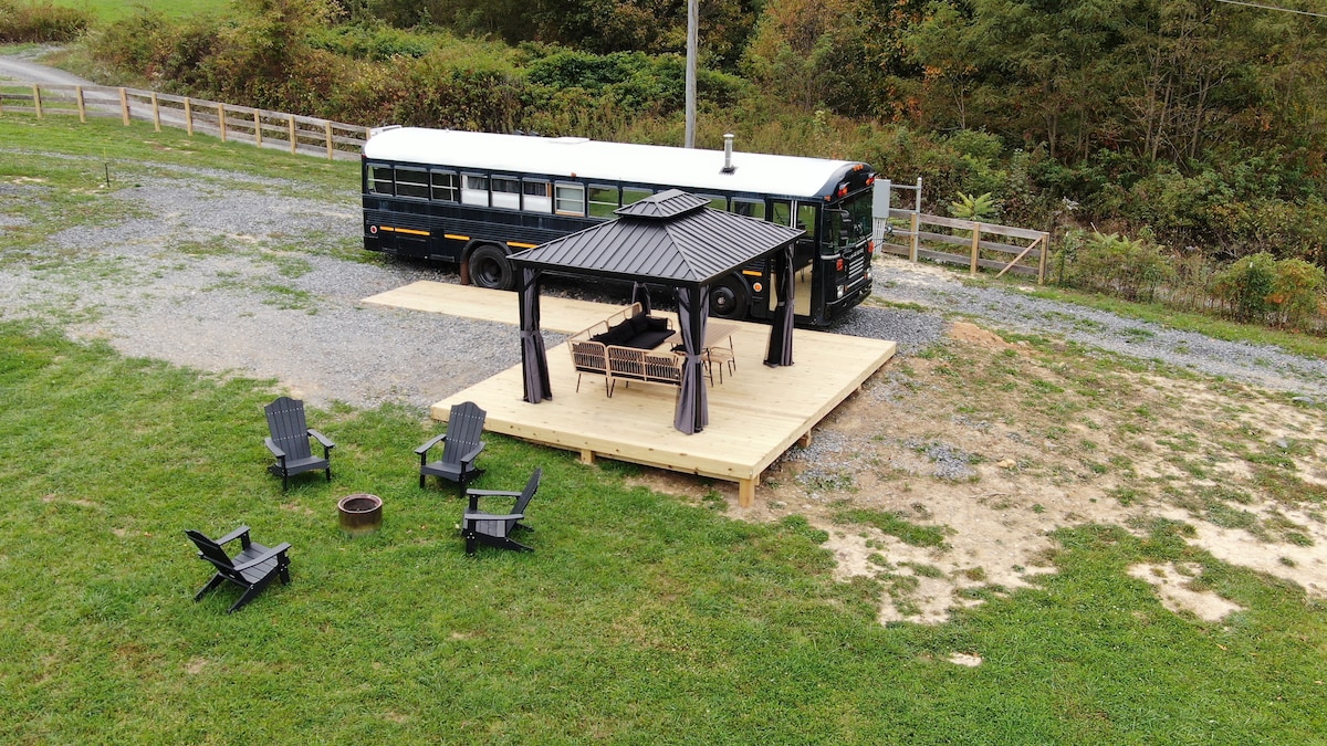 An outdoor area features a wooden deck with a shaded seating arrangement under a gazebo. Surrounding the deck are several black adirondack chairs and a fire pit. A school bus is parked nearby, with grassy fields and trees in the background.