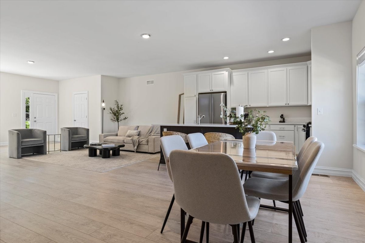 An open concept living area features light hardwood flooring and large windows allowing natural light to fill the space. A rectangular dining table with upholstered chairs sits next to a cozy seating arrangement, which includes a modern sofa and two armchairs. A well-equipped kitchen is visible in the background.