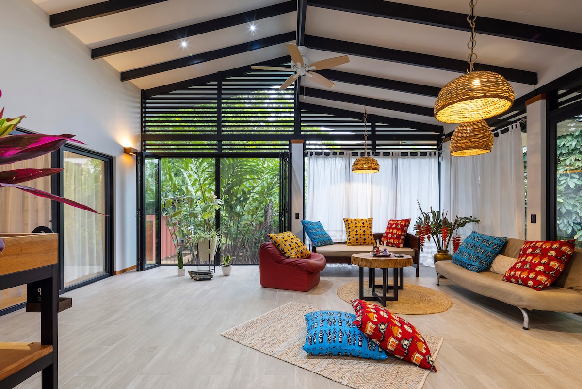 A welcoming living area features high ceilings with exposed beams. A large circular table sits in the center, surrounded by a mix of colorful cushions on various seating options. Large glass doors open to a lush garden view, enhancing natural light and ventilation.