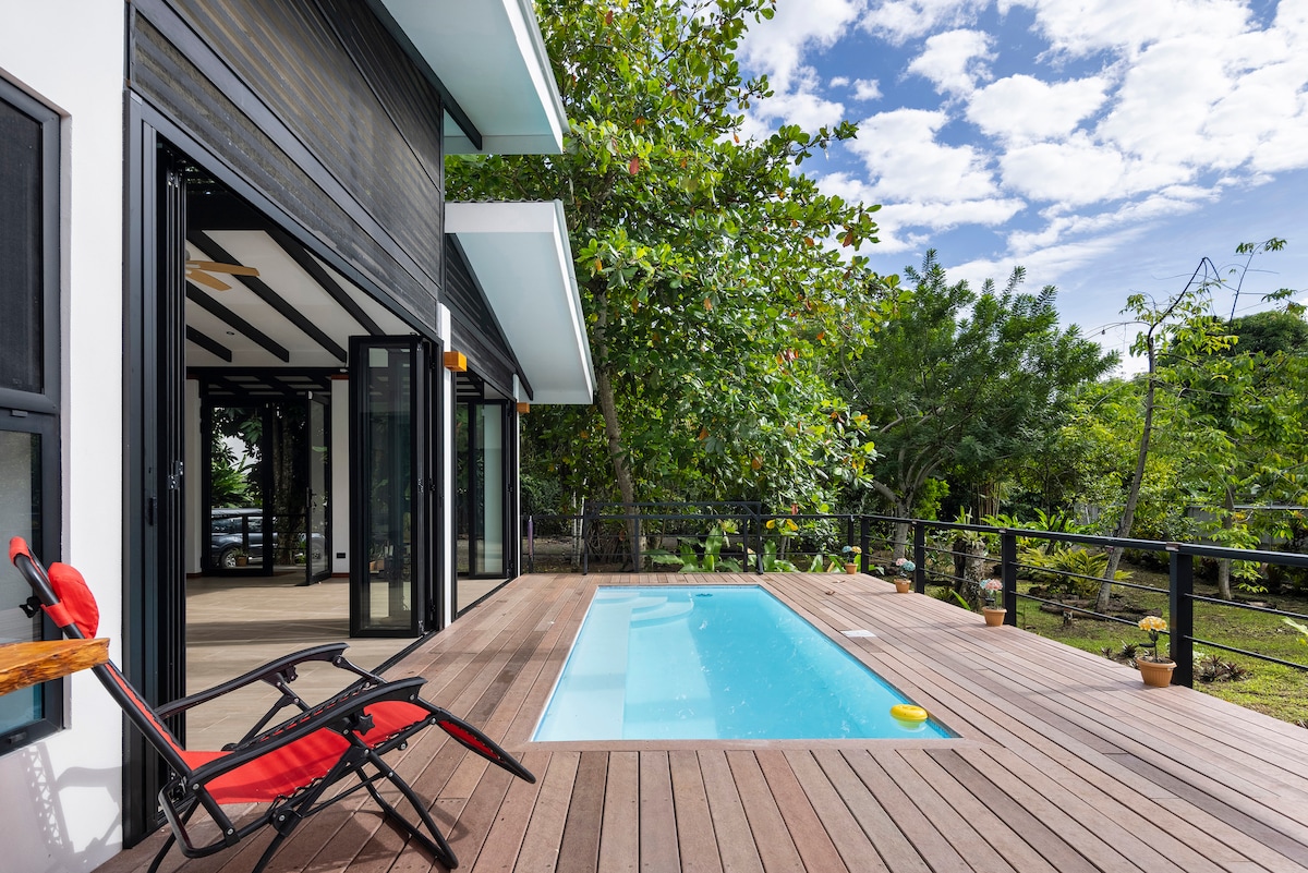 A small swimming pool is situated on a wooden deck, surrounded by lush greenery. Large glass doors open to connect indoor and outdoor spaces, allowing natural light to fill the area. A red lounge chair rests on the deck, inviting relaxation.