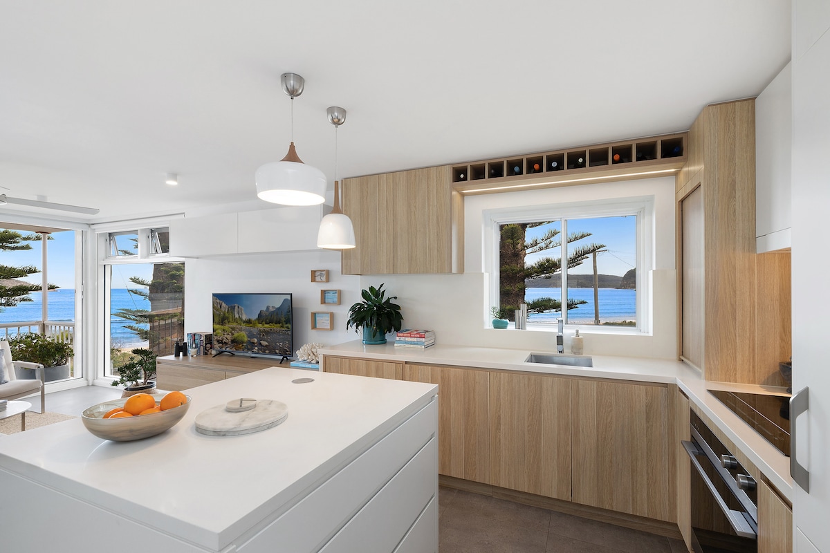 A modern kitchen area features light wood cabinetry and a large island with a marble surface. Natural light floods the space through a window that overlooks the ocean. An attractive bowl of oranges is placed on the island, adding a touch of color.