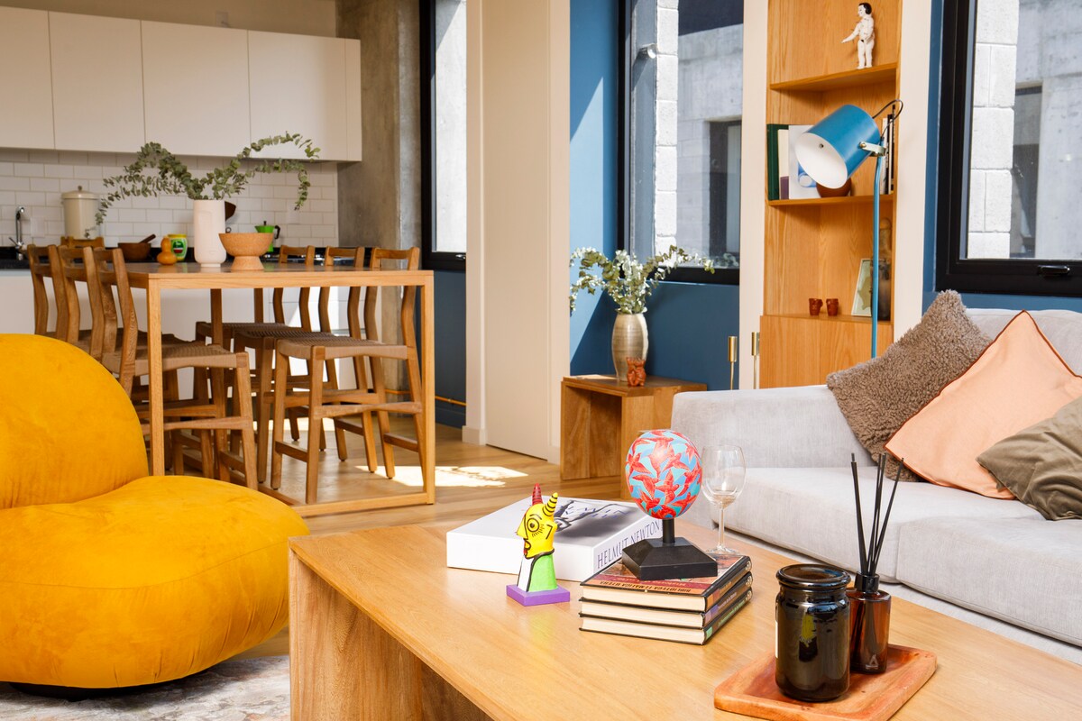 A modern living area showcases a light gray sofa adorned with textured cushions, complemented by a wooden coffee table topped with decorative objects. A bright yellow bean bag adds a pop of color. In the background, a dining area with a light wood table and chairs is visible, alongside a well-lit kitchen.