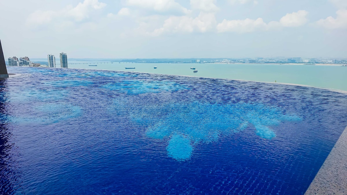 An infinity swimming pool extends towards the horizon, featuring a shimmering blue surface that blends with the sea. The scene reveals a serene sky, complemented by distant high-rise buildings and boats along the water's edge, emphasizing relaxation and tranquility.