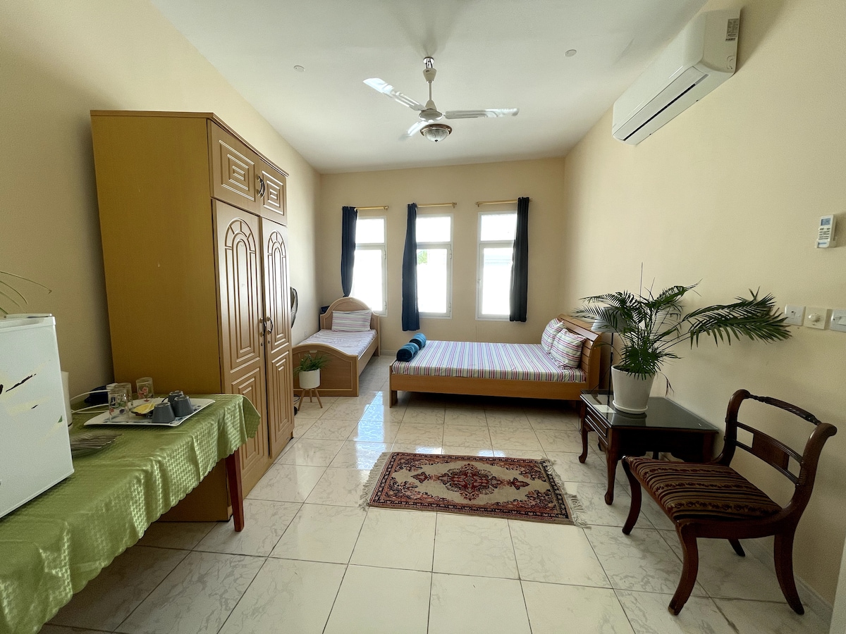 A bright guest room features two comfortable beds, one with a patterned covering. A wardrobe stands against the wall, next to a small sitting area with a chair and a table. Natural light enters through three windows, enhancing the space's openness.