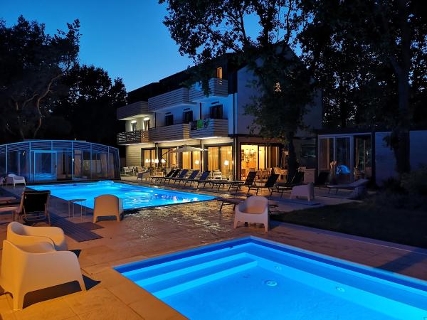 A modern building features large glass windows reflecting warm lighting from within. Two pools, one larger and one smaller, are illuminated in blue, surrounded by lounge chairs. The setting is framed by trees, enhancing a peaceful outdoor atmosphere.