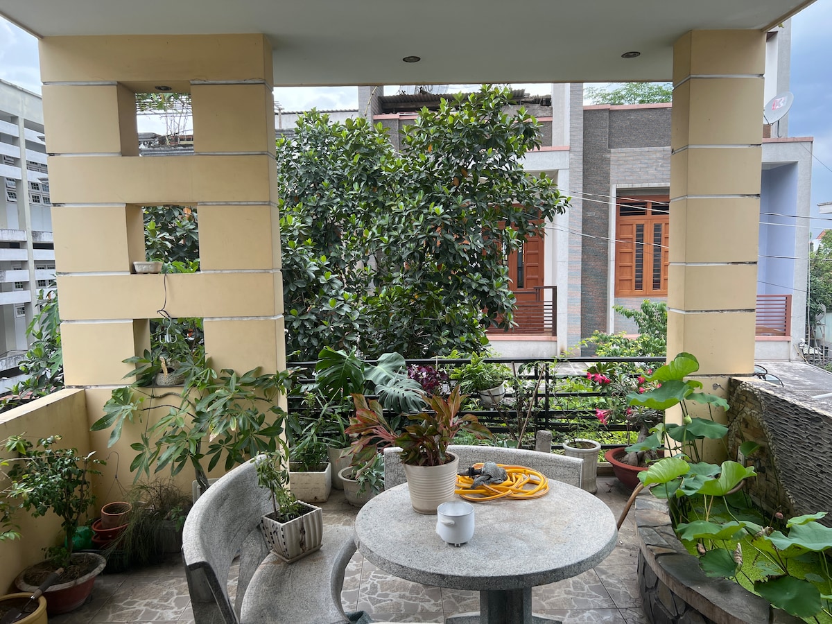 A covered terrace is surrounded by lush greenery in various planters. A round table is positioned with a textured surface, accompanied by two rounded chairs. A potted plant features a modern, minimalistic design, enhancing the outdoor space.
