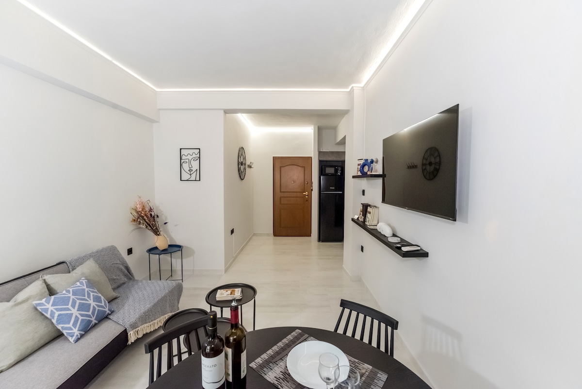A modern living space features a sectional sofa with accent cushions, complemented by a round dining table set for two. A wall-mounted television is visible, alongside minimalist shelves displaying decor. Soft lighting enhances the white walls, creating a bright and open ambiance.