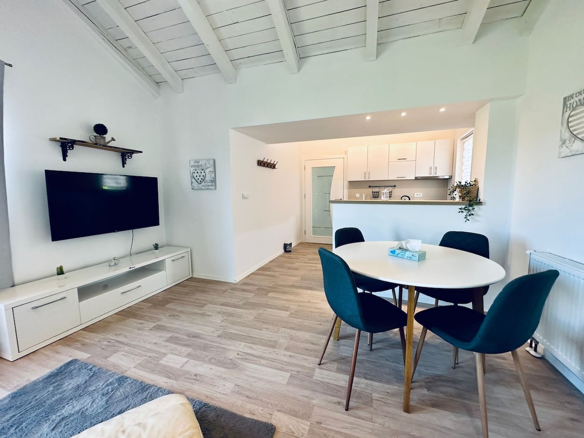 A light-filled living space features a flat-screen TV mounted on the wall and a round dining table surrounded by four chairs. The open kitchen area, equipped with modern appliances, is visible in the background. White beams accent the high ceiling, enhancing the airy feel of the room.