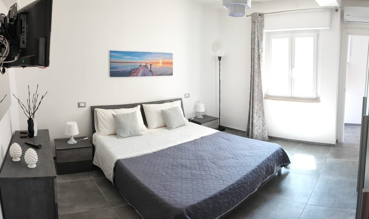 A spacious bedroom features a large bed with neutral bedding and decorative pillows. Two bedside tables hold lamps. A flat-screen television is mounted on the wall. Natural light enters through a window with sheer curtains, while a floor lamp in the corner adds additional illumination.