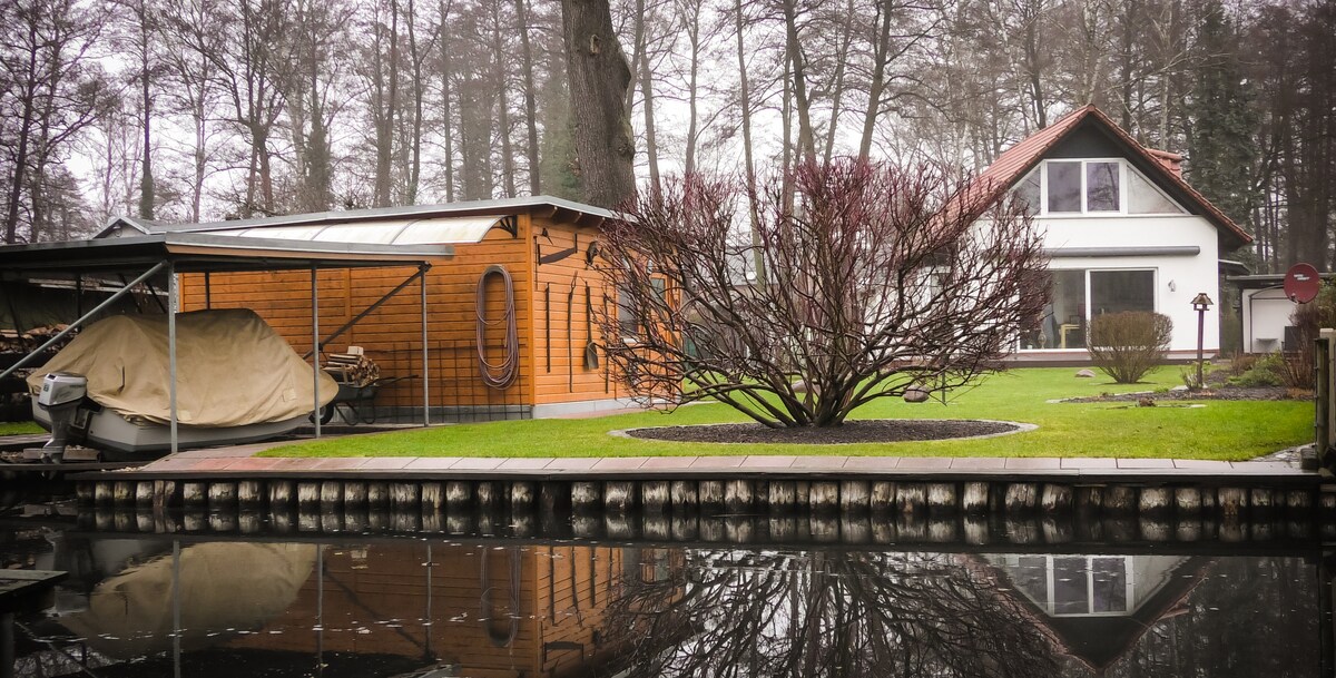 A serene view of the property alongside the river, highlighted by a wooden boathouse partially covered. A vibrant tree with bare branches stands prominently in the garden, which is bordered by carefully maintained grass. The house exhibits a modern architectural style with large windows.