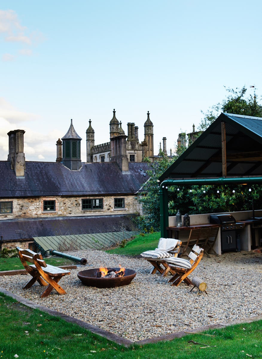 A stone courtyard is depicted with a fire pit surrounded by wooden chairs. A covered area, featuring a grill and outdoor kitchen, is positioned nearby. The historic facade of Narrow Water Castle can be seen in the background, framed by greenery and a clear sky.