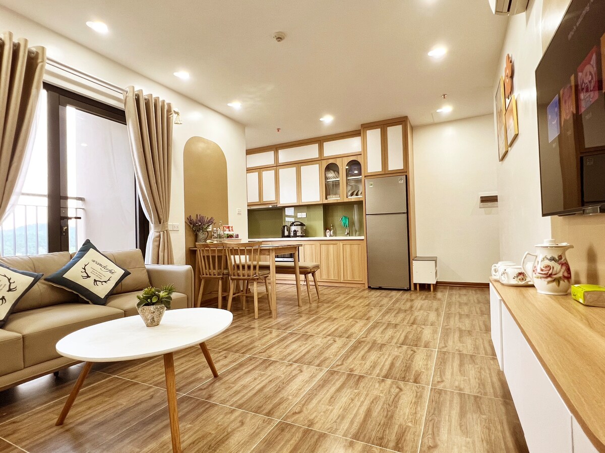 A modern living space features a light-coloured sofa arranged around a circular coffee table. In the background, a kitchen area is visible with wood-toned cabinetry, a refrigerator, and appliances. Natural light enters through large balcony doors, contributing to a bright interior.