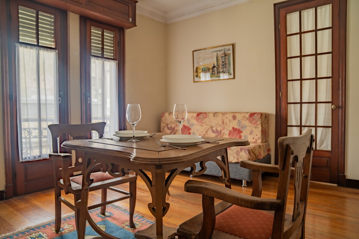 A dining area features a wooden table set for two, accompanied by two vintage-style chairs. A floral-patterned sofa is positioned against the wall, with large windows allowing natural light to fill the space. Wooden details in the door and window frames enhance the charm.