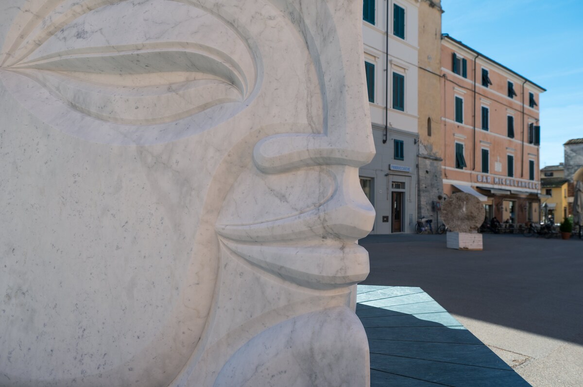 An intricately carved marble sculpture stands prominently in the square, highlighting smooth curves and facial features. In the background, a charming building with multiple windows and pastel-colored façades complements the artistic environment, while a large, rounded stone adds to the outdoor space.