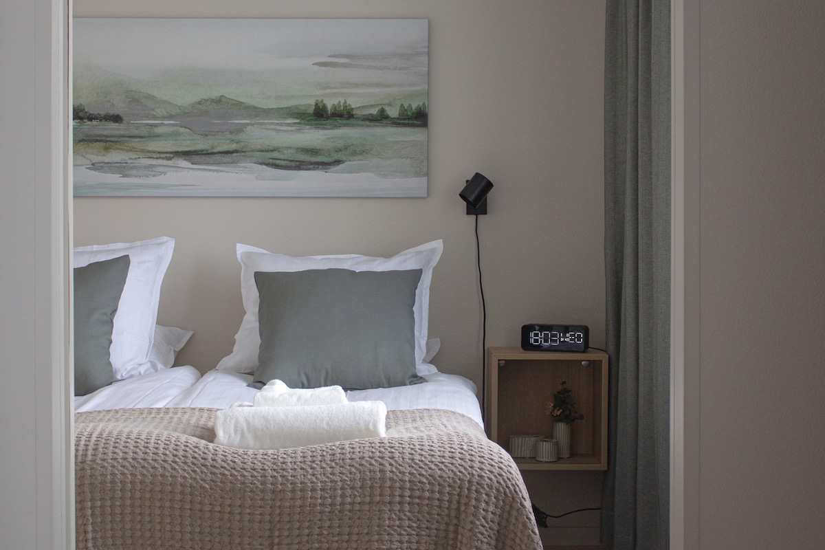 The bedroom features two twin beds with light linens and green accent pillows. A simple artwork depicting a landscape hangs above the beds. A nightstand with a clock and a small plant is located next to the bed, providing essential bedside accessories.