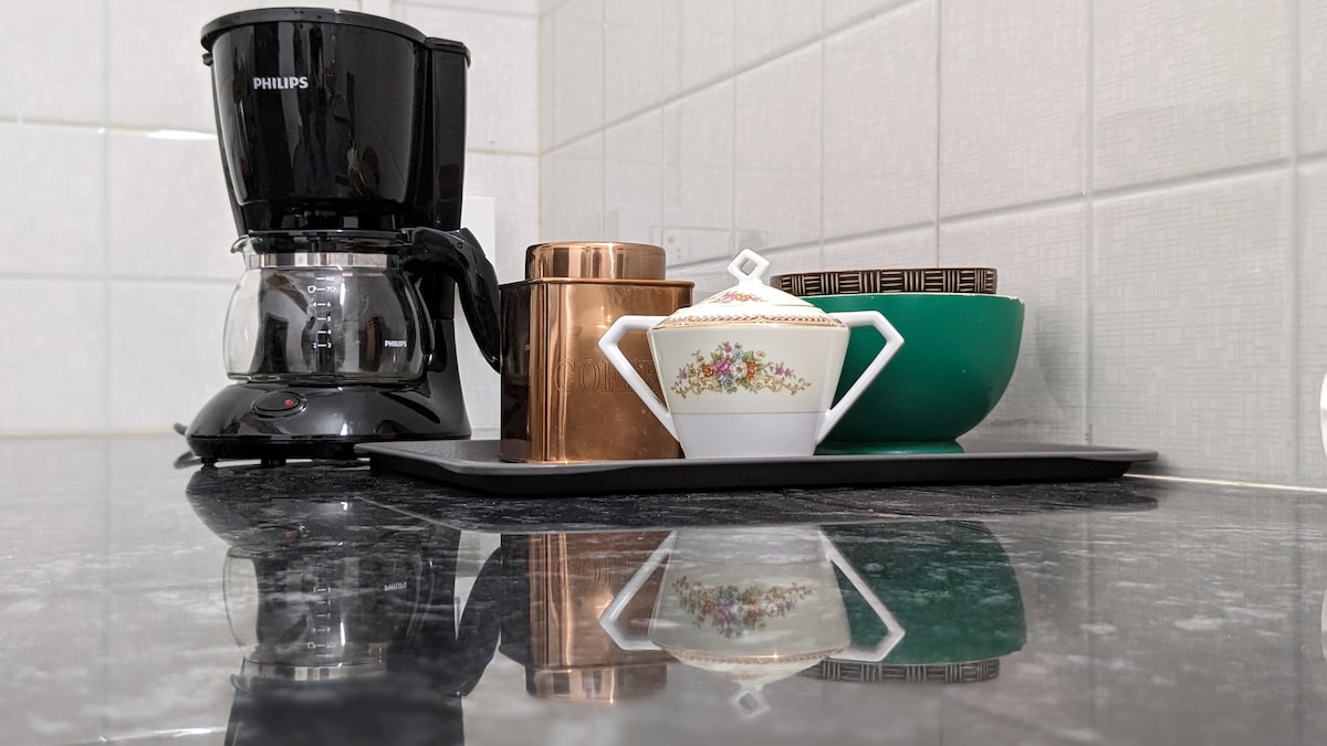 A coffee maker and three containers are arranged on a dark countertop. The coffee maker is black, and a polished copper canister sits beside a colorful green bowl and a white ceramic sugar bowl with floral detailing. Reflections on the surface enhance the visual appeal.