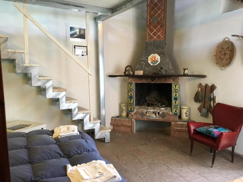 A cozy room features a spacious bed with white linen, complemented by a red armchair nearby. The rustic stone fireplace is embellished with colorful tile accents. A wooden staircase leads to an upper level, while framed photographs adorn the walls.