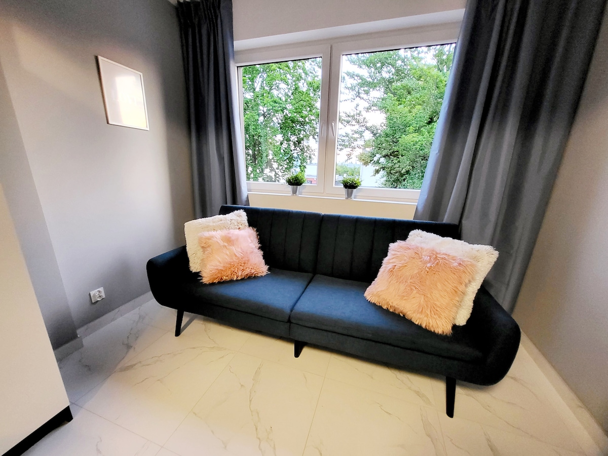 A modern seating area is showcased with a dark sofa adorned with two plush, light-colored pillows. Large windows allow natural light to fill the space, framed by floor-to-ceiling curtains, while greenery is visible outside.