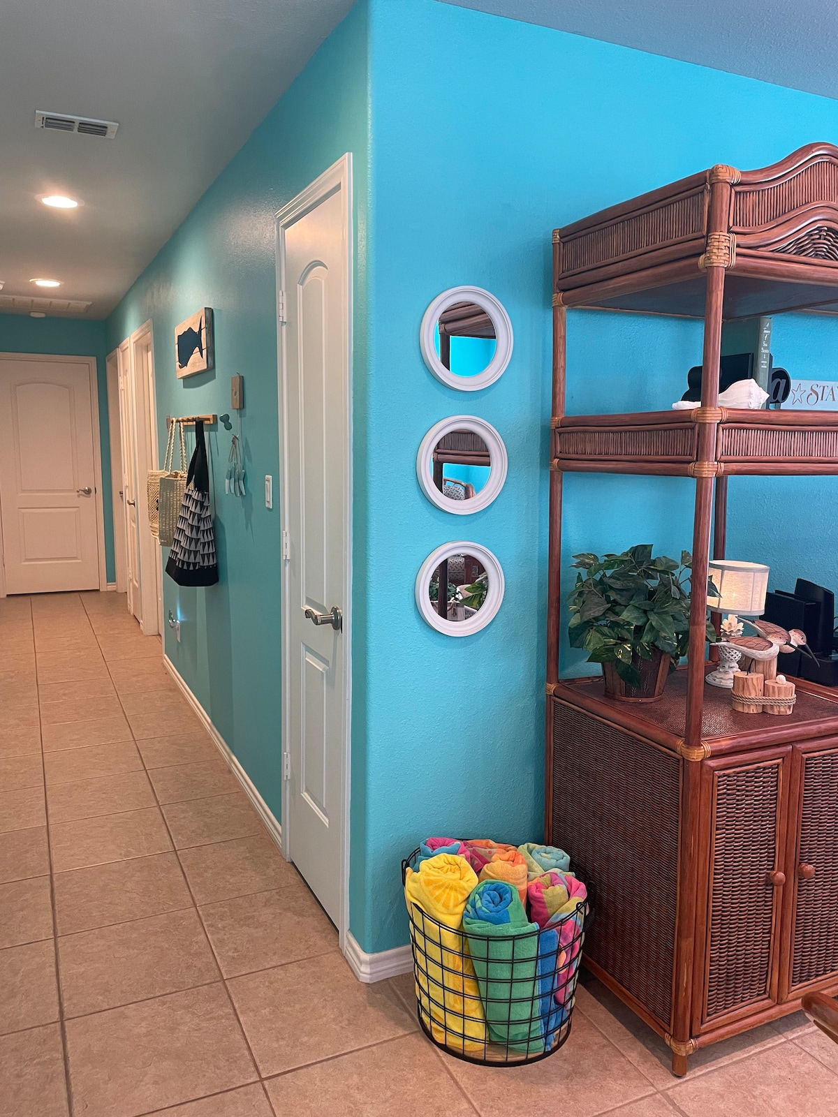 A bright hallway is characterized by turquoise walls and tiled flooring. Three circular mirrors are mounted on one wall, while a woven storage unit displays plants and decorative items. A colorful basket holds neatly rolled towels, adding a cheerful touch to the space.