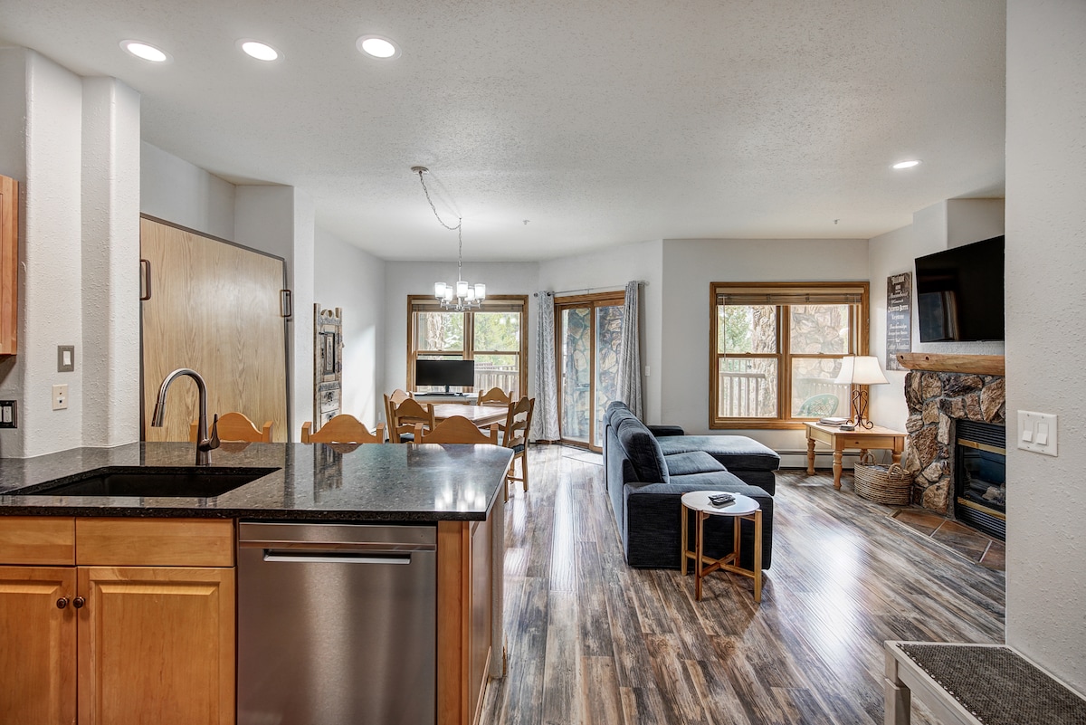 An open-concept living area is visible, featuring a kitchen with wooden cabinetry and granite countertops. A dining area with a wooden table and multiple chairs is adjacent to a cozy seating arrangement by a stone fireplace. Large windows bring in natural light, showcasing views of the outdoors.