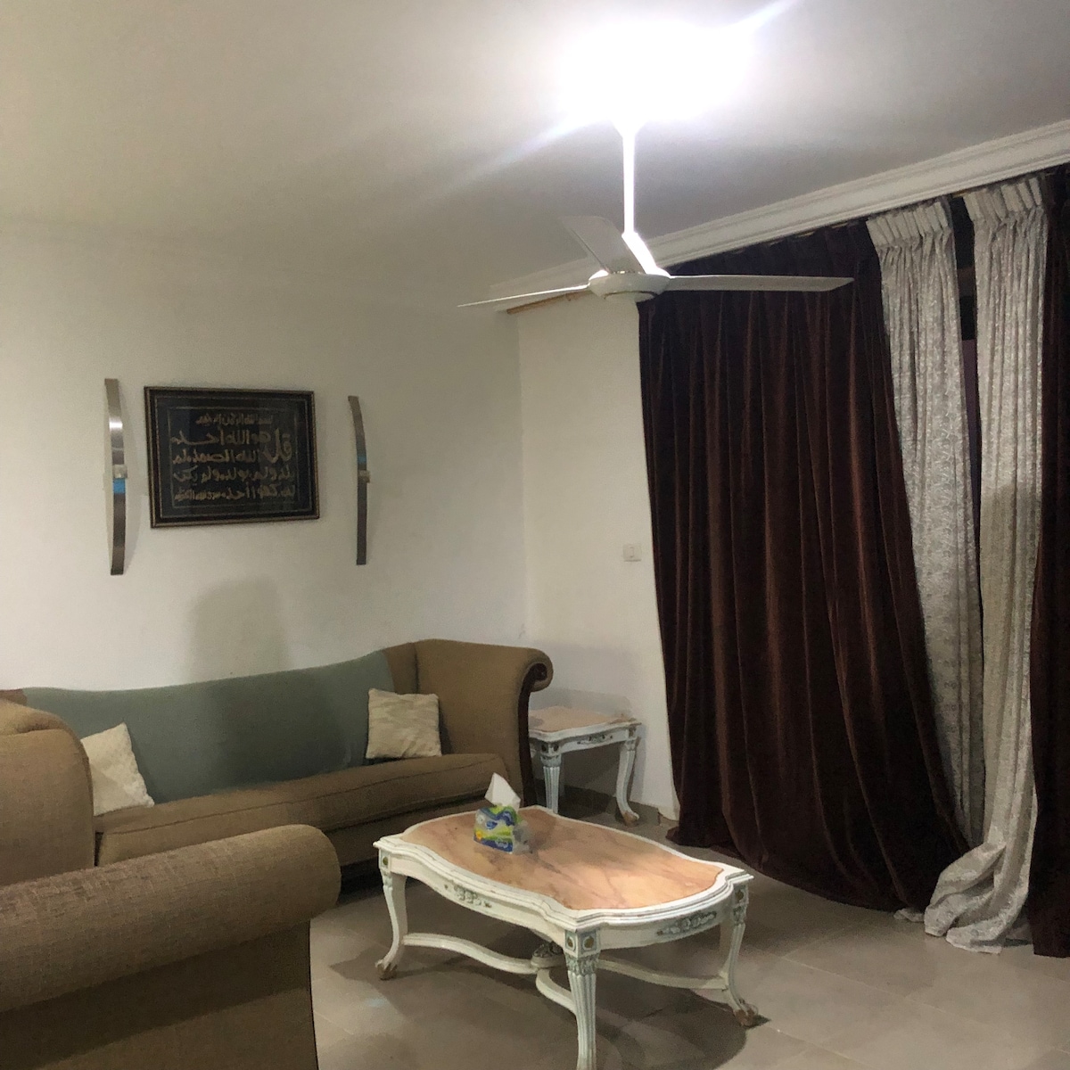 A living area is depicted with a neutral-colored sofa adorned with decorative pillows. A light-colored coffee table sits at the center, topped with a small decorative item. Dark curtains frame the window, while a ceiling fan provides overhead airflow.