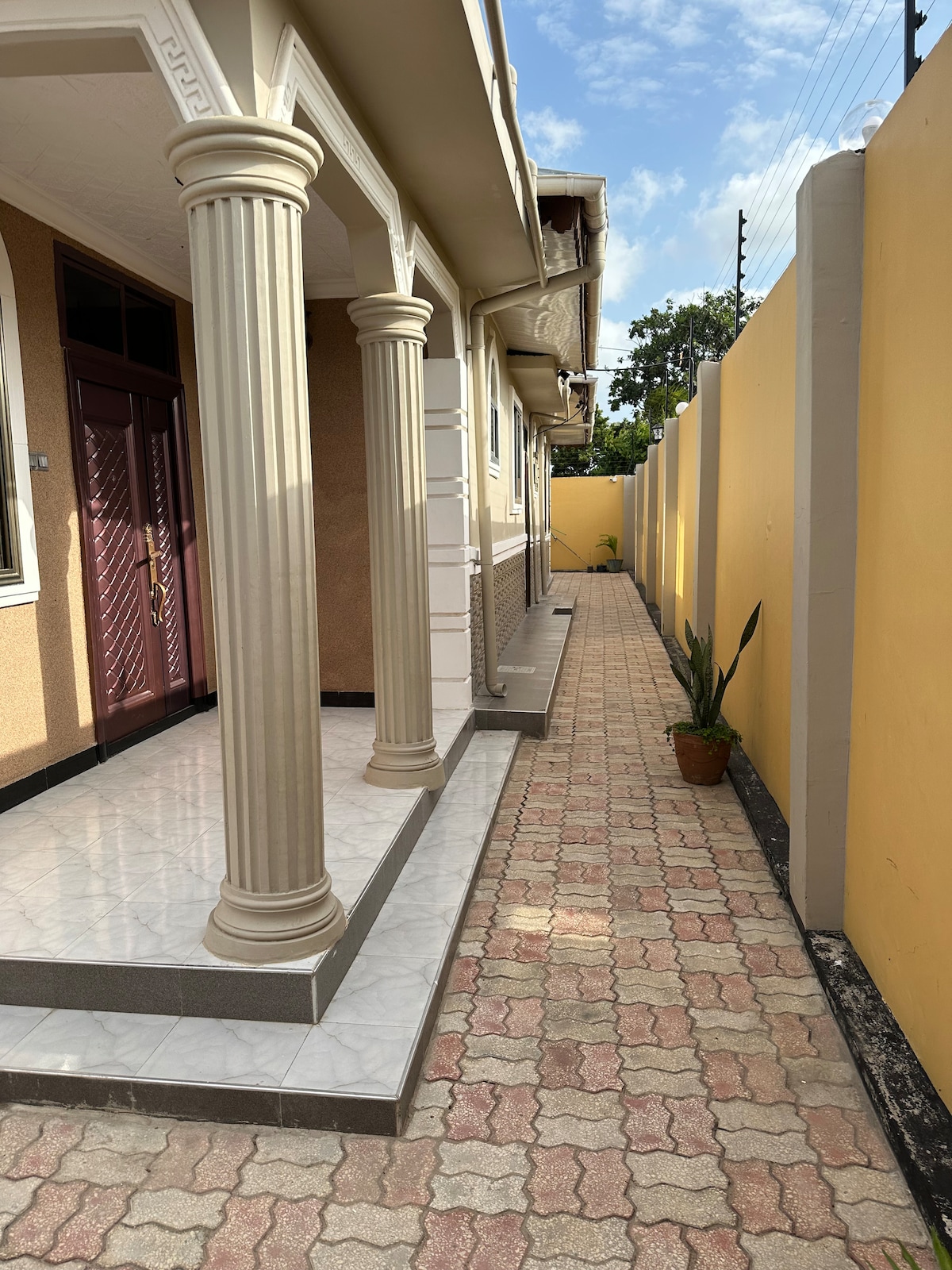 A pathway lined with patterned tiles extends alongside the building, bordered by yellow walls. The entrance features elegant columns supporting the overhang, while a potted plant adds a touch of greenery near the edge of the path.