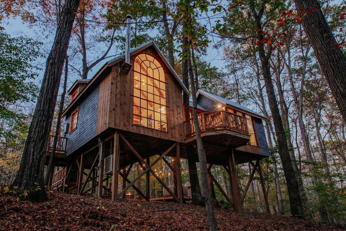 The treehouse is elevated on stilts, surrounded by tall trees and autumn foliage. Large windows dominate the front, allowing natural light to illuminate the interior. A spacious deck extends from one side, featuring a seating area. The serene setting offers a sense of retreat in nature.