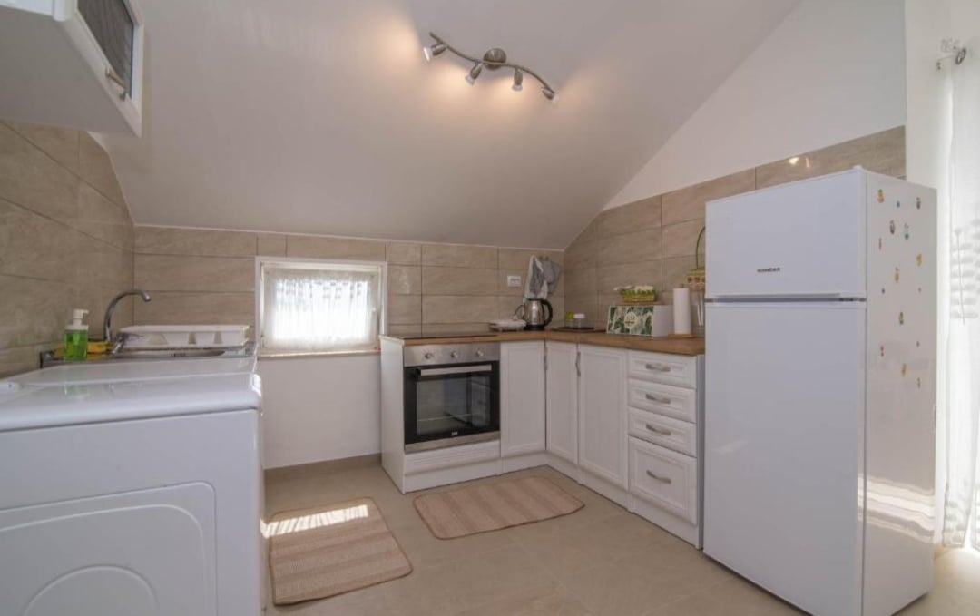 A functional kitchen is equipped with essential appliances, including a washing machine, oven, and refrigerator. Clean lines and ample counter space enhance the area's usability. Natural light filters through a small window, creating a bright atmosphere. Soft rugs add warmth underfoot.