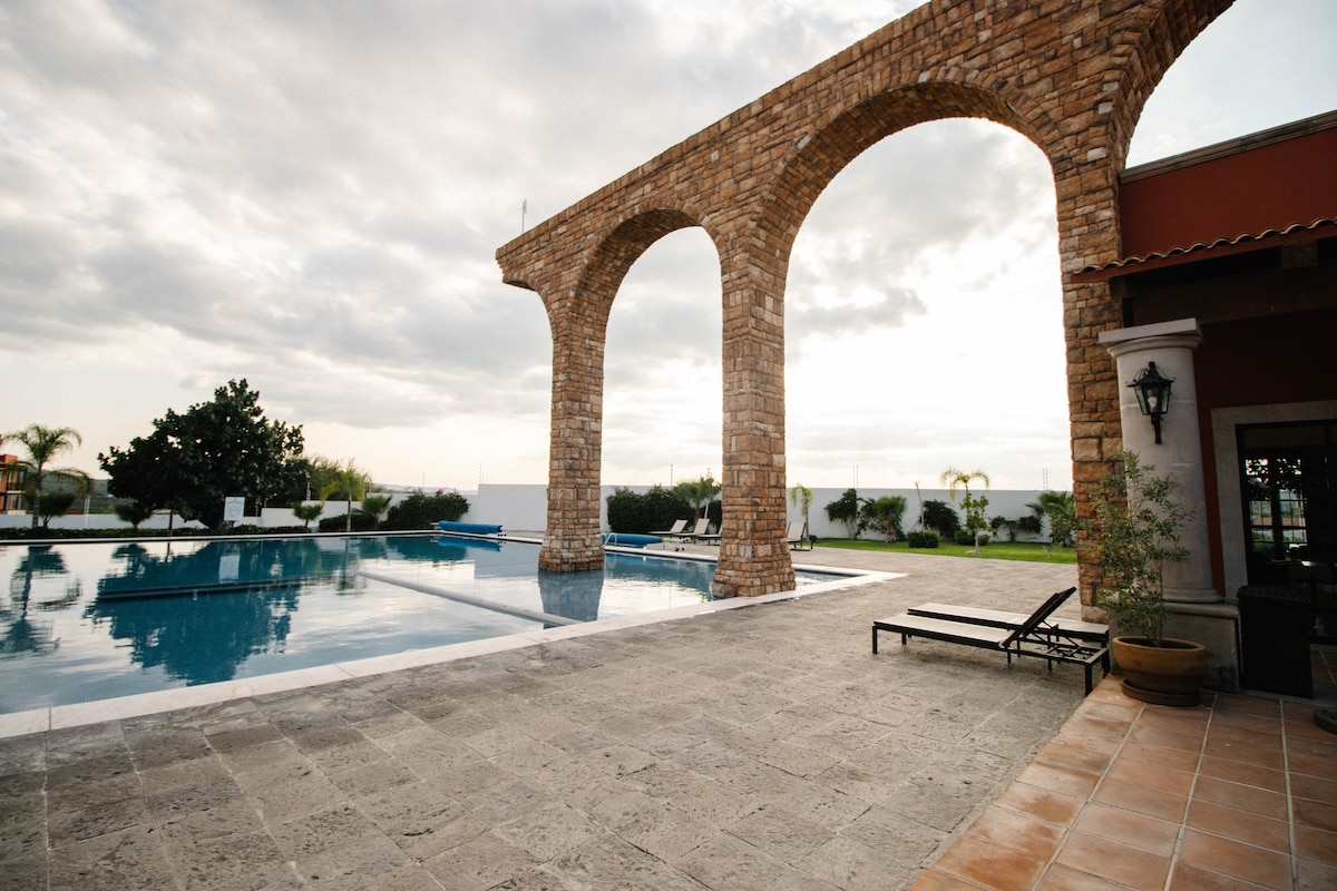 An expansive outdoor area features a large swimming pool, with a striking stone archway framed against the sky. Lounge chairs sit nearby on a patterned stone surface, providing a space to relax. Lush greenery surrounds the pool area, enhancing the tranquil atmosphere.