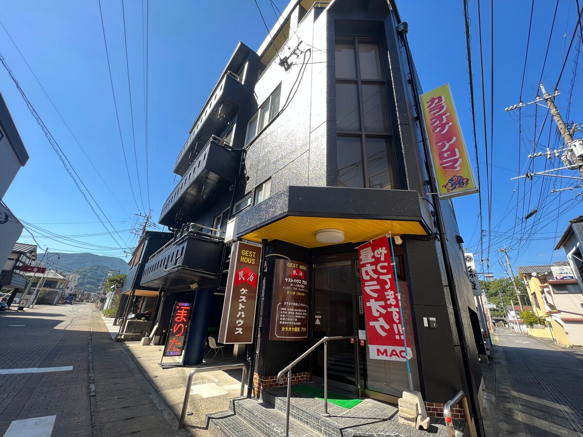 The exterior of a modern guesthouse is showcased, featuring a sleek black facade and multiple levels. A welcoming entrance with steps leads to the main door, where signs display information regarding the accommodations. Surrounding power lines and mountains create a scenic backdrop.