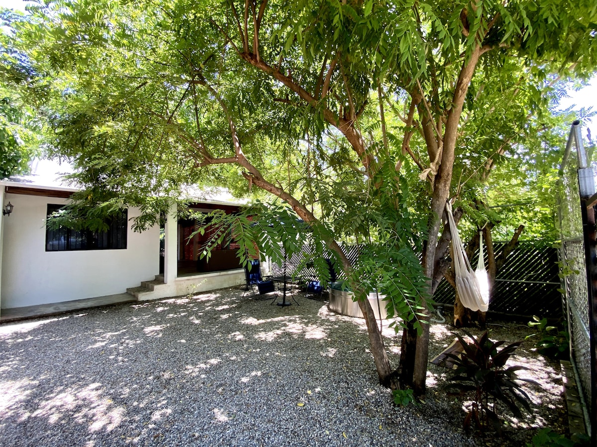 A spacious outdoor area featuring a gravel surface is shaded by lush trees. A hammock is gently swaying between the branches, while a small table and chairs are positioned nearby. The house is visible in the background, surrounded by greenery.