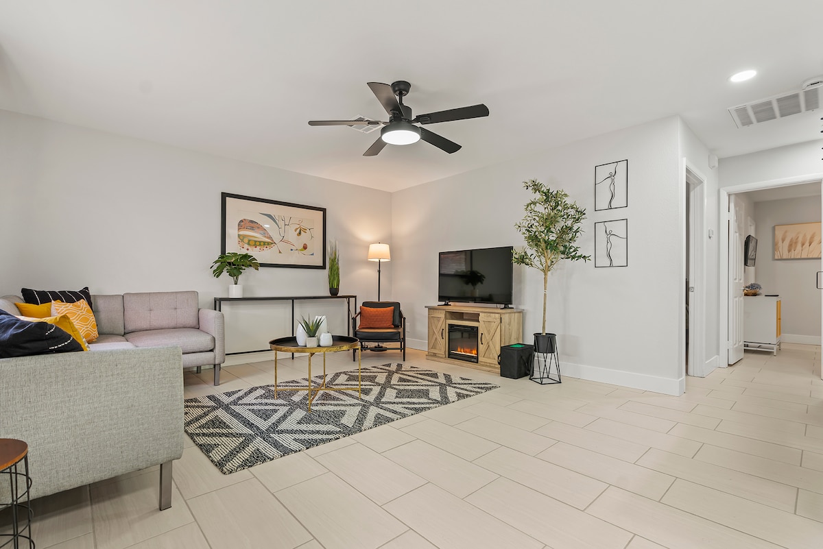 The open-concept living area showcases a comfortable sofa and accent chair, complemented by a decorative rug and a coffee table. A television is mounted on the wall, with a faux fireplace nearby. Natural light enhances the spacious atmosphere, promoting relaxation.