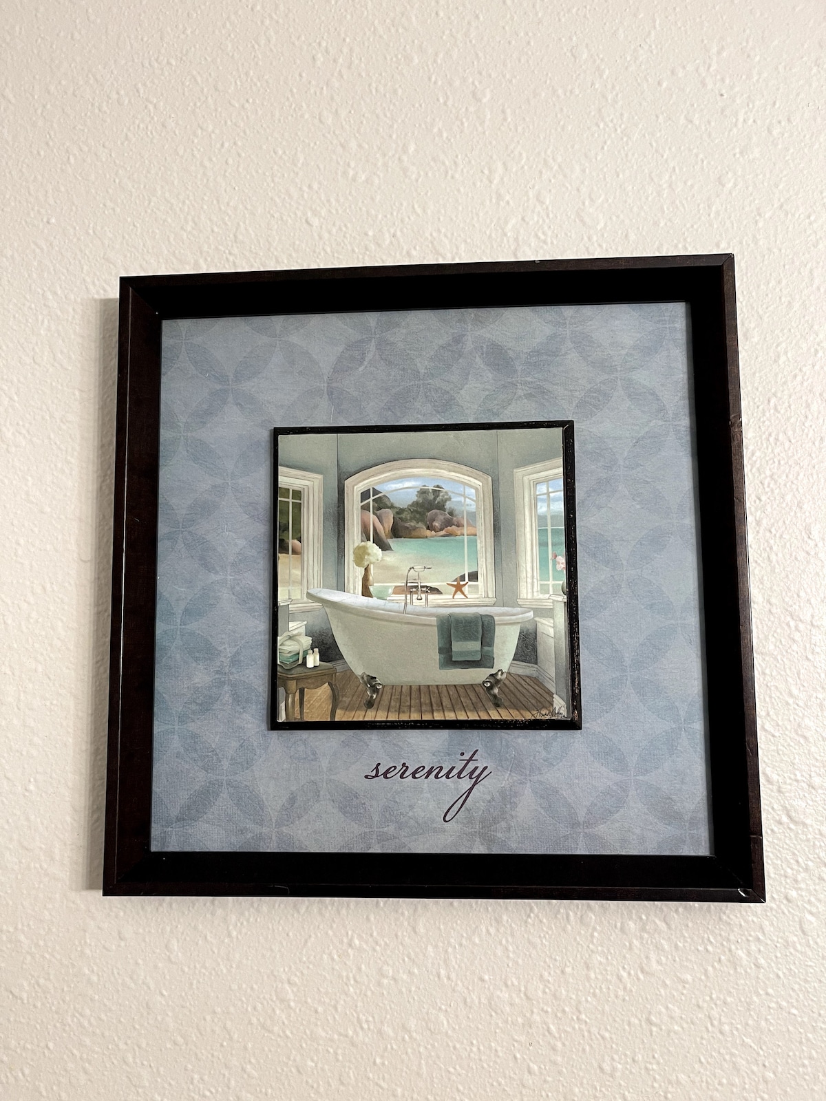 A framed picture hangs on a textured wall, depicting a bathroom scene. The image features a freestanding bathtub positioned under a large window, with soft natural light illuminating the space. The word 'serenity' is artistically displayed beneath the image on a subtly patterned background.