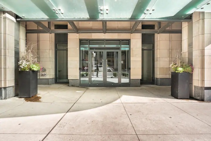 An entrance to the building is shown, featuring a sleek glass canopy overhead. Large planters with greenery are positioned on either side of the entrance, and a smooth concrete pathway leads towards the doors, allowing natural light to illuminate the area.