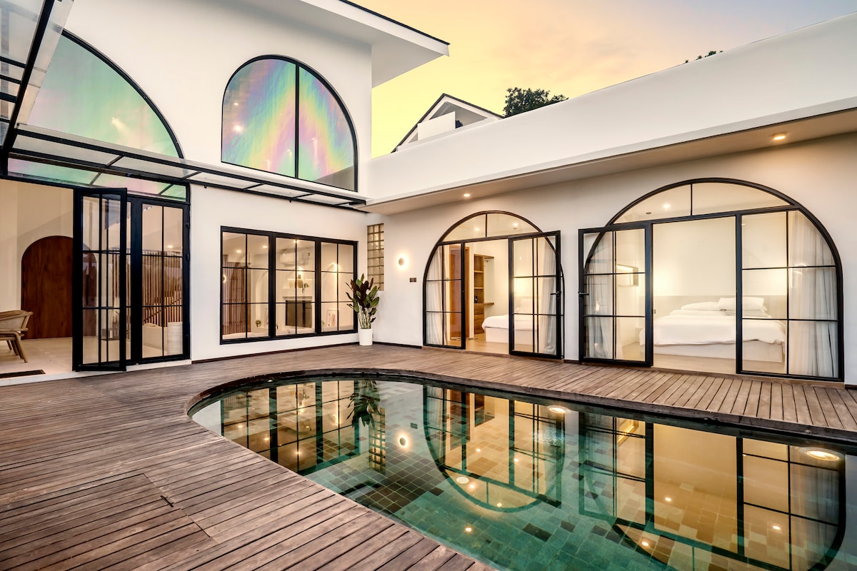 An outdoor view presents an inviting private pool surrounded by wooden decking. Arch glass doors lead to an elegant living space and two cozy bedrooms, showcasing the sleek design of the villa at sunset. The ambiance reflects a tranquil retreat atmosphere.